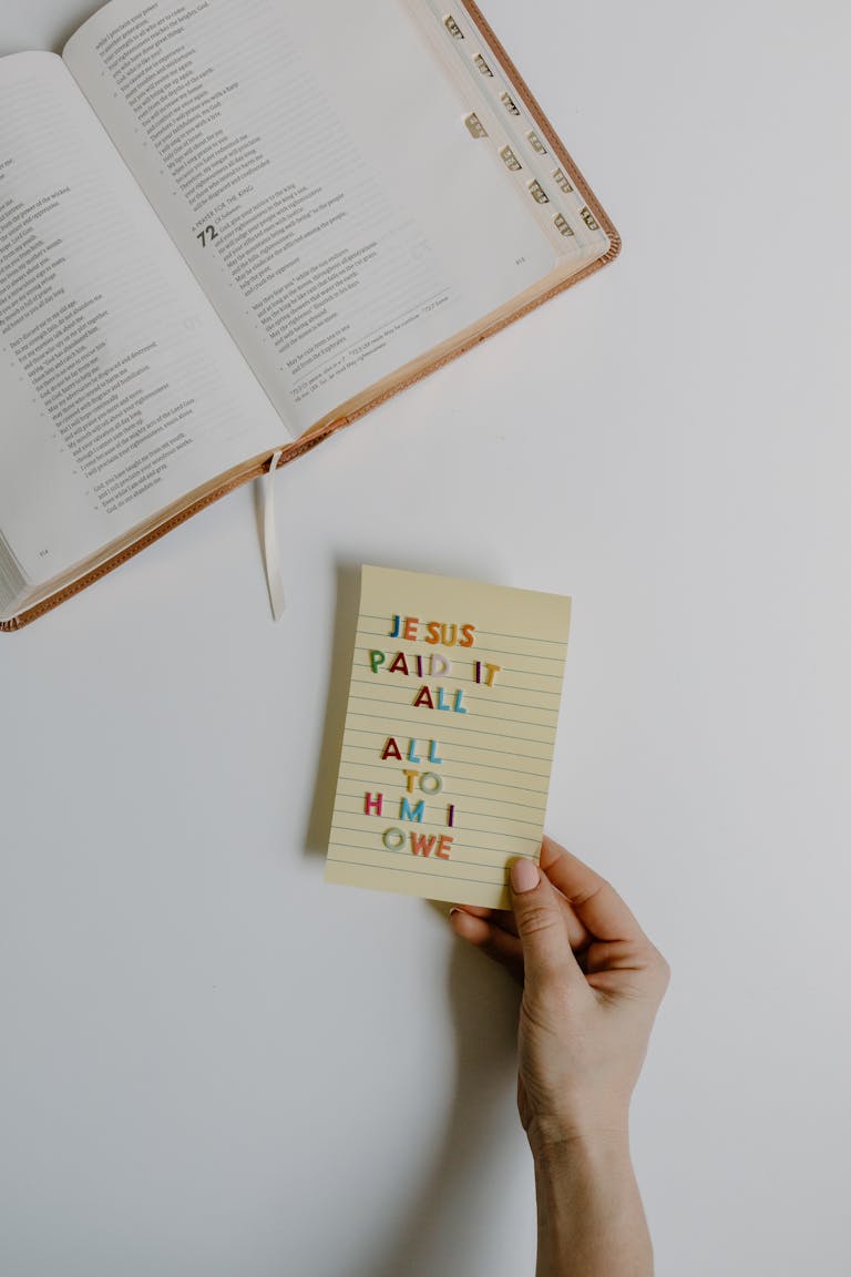 Hand holding a note with a Bible verse beside an open Holy Bible, symbolizing faith.